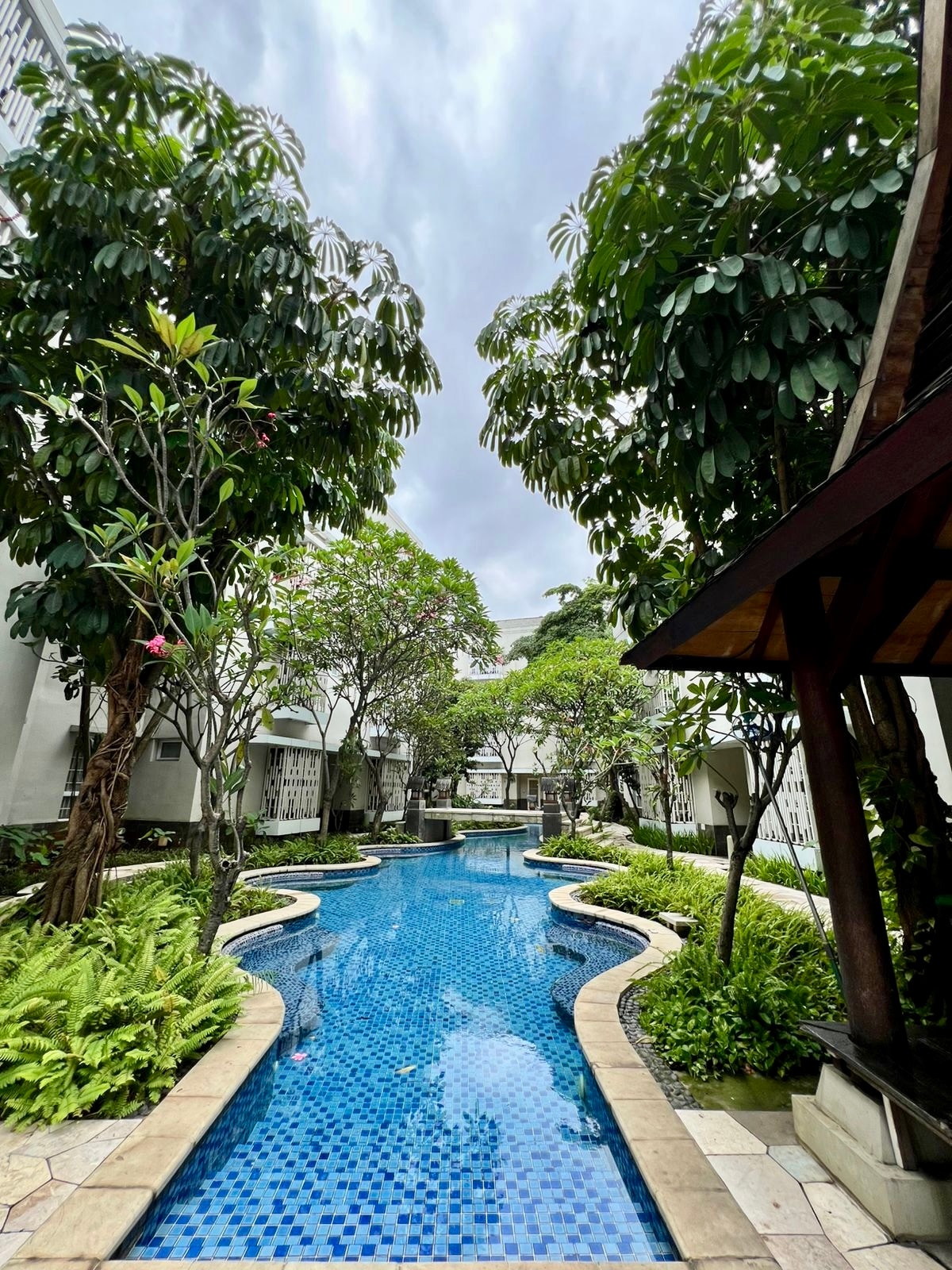 A lagoon-like swimming pool winds through lush greenery, featuring vibrant blue tiles that reflect the sky. Surrounded by tropical plants and trees, the tranquil setting enhances the sense of relaxation. Pathways along the pool lead to shaded sitting areas amidst the serene landscape.