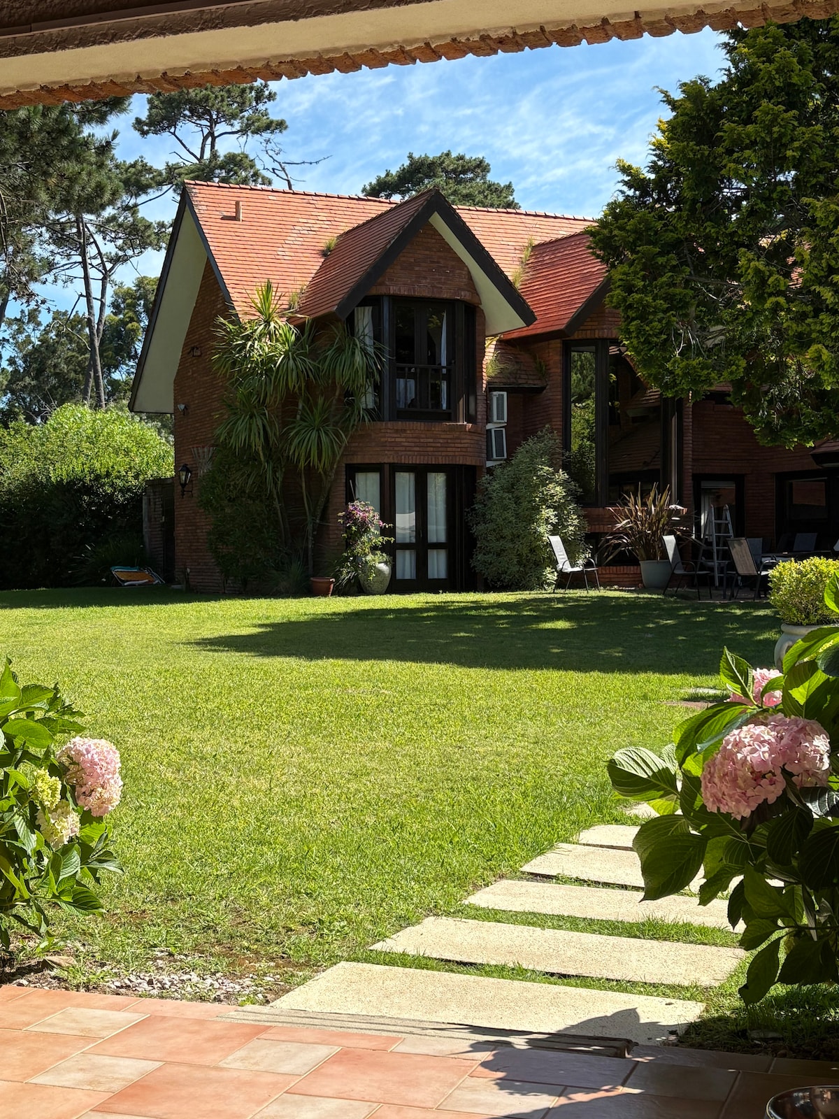 An exterior view of the home showcases a brick structure surrounded by a lush green garden. A pathway leads to the entrance, flanked by blooming hydrangeas. Large windows reflect natural light, while trees provide shade, enhancing the tranquil atmosphere of the property.