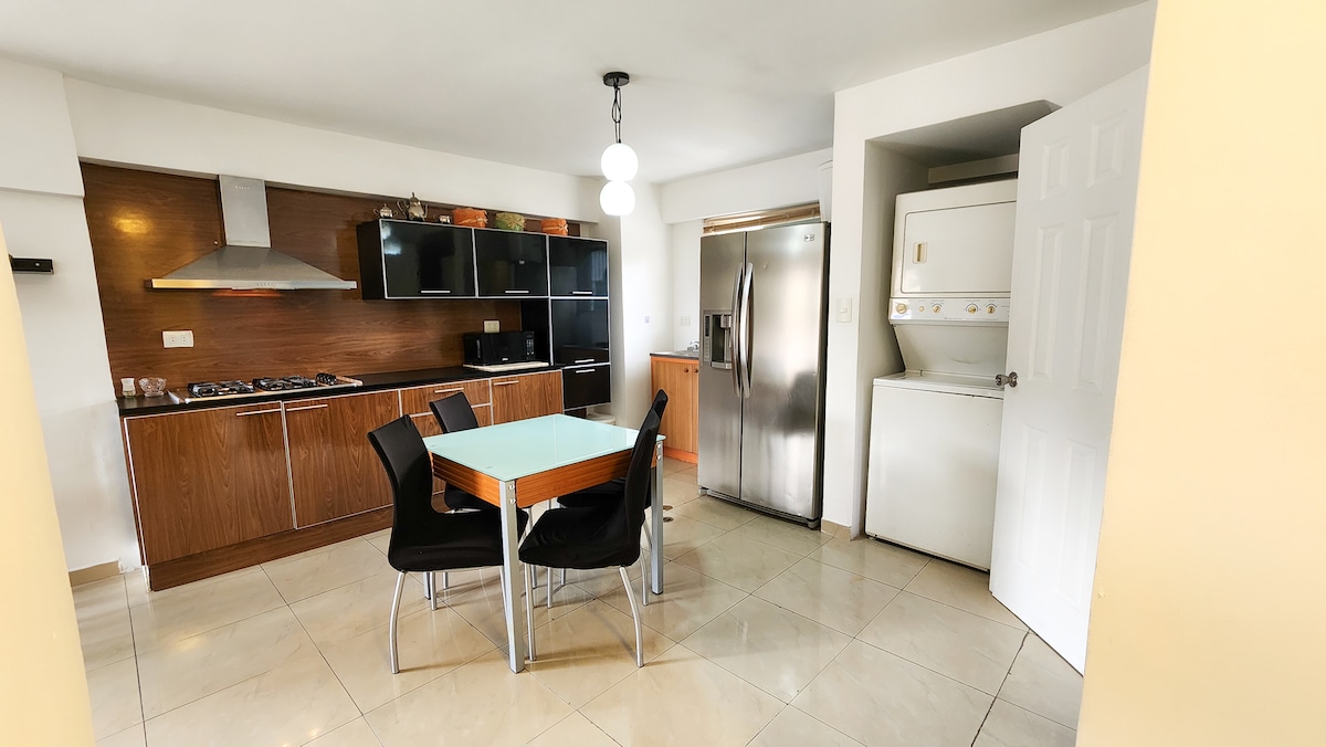 A modern kitchen area features a dining table with four black chairs. The space includes a stainless steel refrigerator, a gas stove with an overhead exhaust, and built-in cabinetry for storage. A washing machine is situated in the corner, enhancing functional convenience.