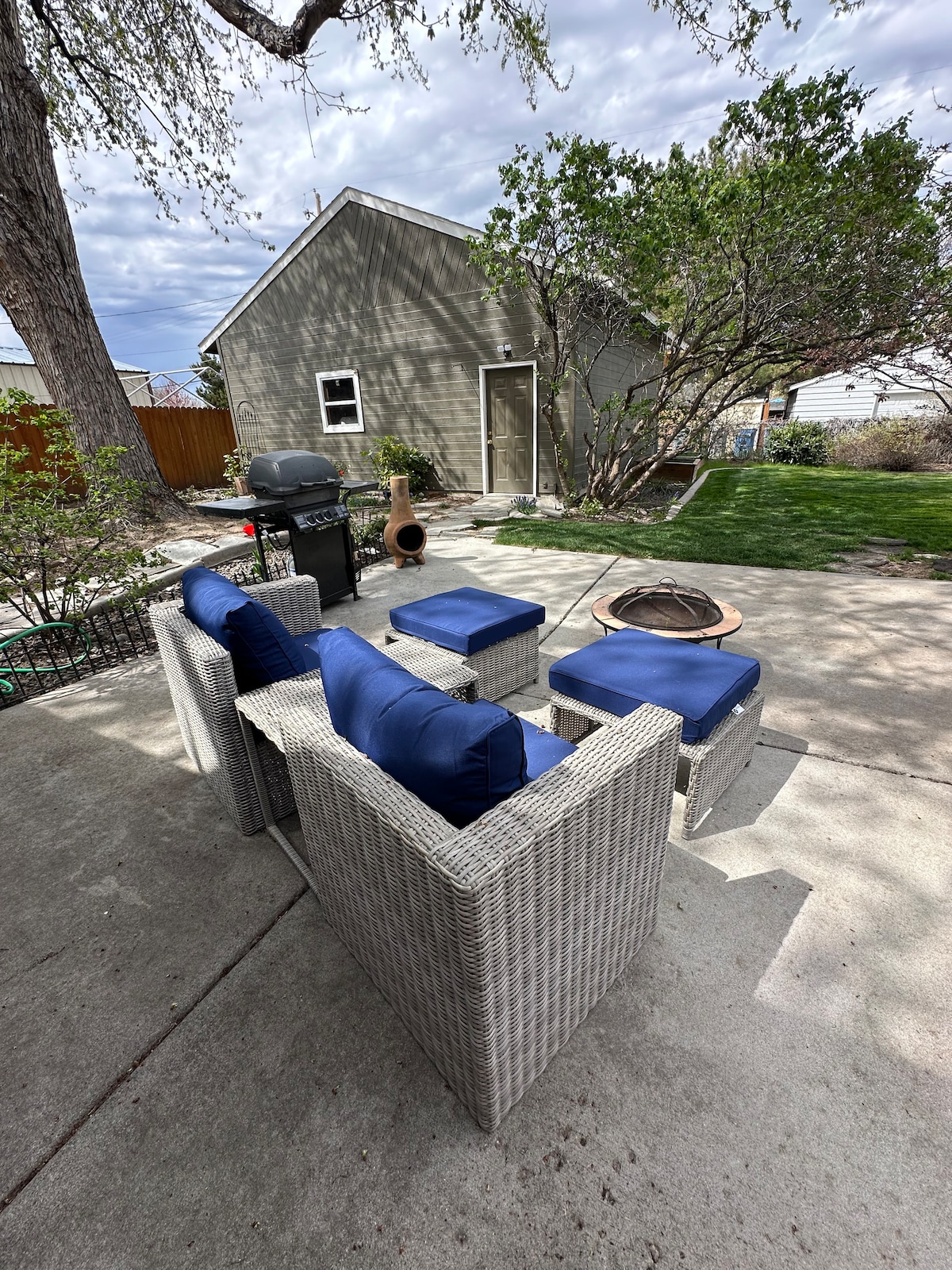 A small outdoor patio area features a set of wicker chairs with blue cushions arranged around a fire pit. A gas barbecue is positioned nearby, with lush greenery and a fence enhancing the private space. The backyard is well-maintained, providing ample room for relaxation.