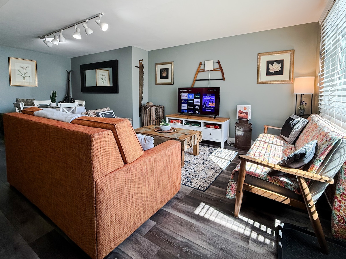 An inviting living room space is showcased, featuring an orange sofa and a floral-patterned wooden bench. A coffee table sits in the center, with a TV mounted on a wall opposite. Natural light streams through blinds, illuminating the room's painted walls and decorative accents.
