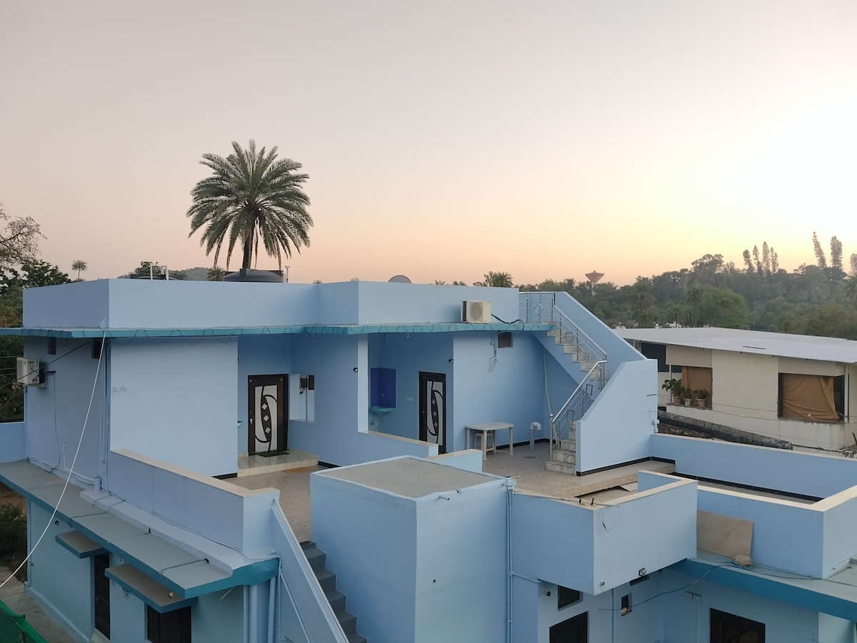 The image captures a multi-level building featuring a light blue exterior under a soft sunset glow. A terrace can be seen with a table set, while staircases and doors add architectural interest. Surrounding palm trees provide a natural setting.