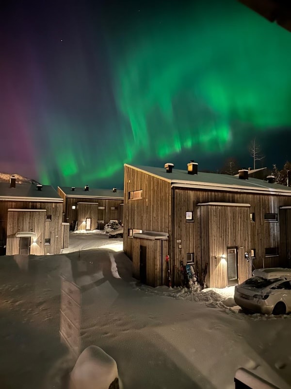 A cluster of modern wooden cabins is set against a snowy landscape under a vibrant display of the Northern Lights. The colorful aurora fills the night sky, creating a serene atmosphere. Snow-covered vehicles are parked nearby, with soft white snow blanketing the ground.