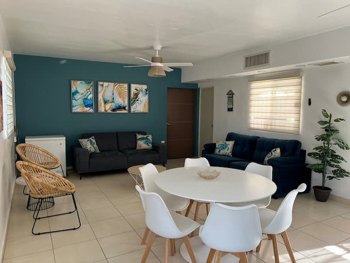 Sala con 2 sillones, uno de ellos es sofá cama con colchón matrimonial, el otro con extensión con espacio suficiente para dormir una persona. Mesa de centro y dos sillas con una mesa pequeña. 1 abanico de techo y AC de tipo central. Cortinas 