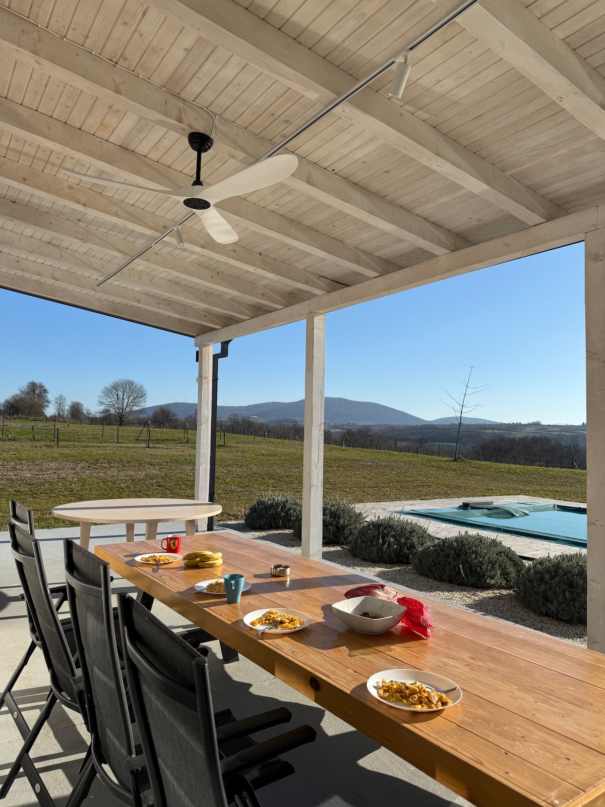 An outdoor dining area is presented with a long wooden table accompanied by several black chairs. Plates filled with food and colorful cups are arranged on the table. A scenic view of rolling hills and an inviting pool can be seen in the background under a clear blue sky.