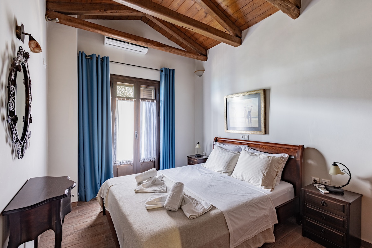 A spacious bedroom features a wooden ceiling with exposed beams. A large bed is dressed in neutral linens, complemented by two bedside tables and lamps. Natural light filters through a window with curtains, offering views of the outdoors.