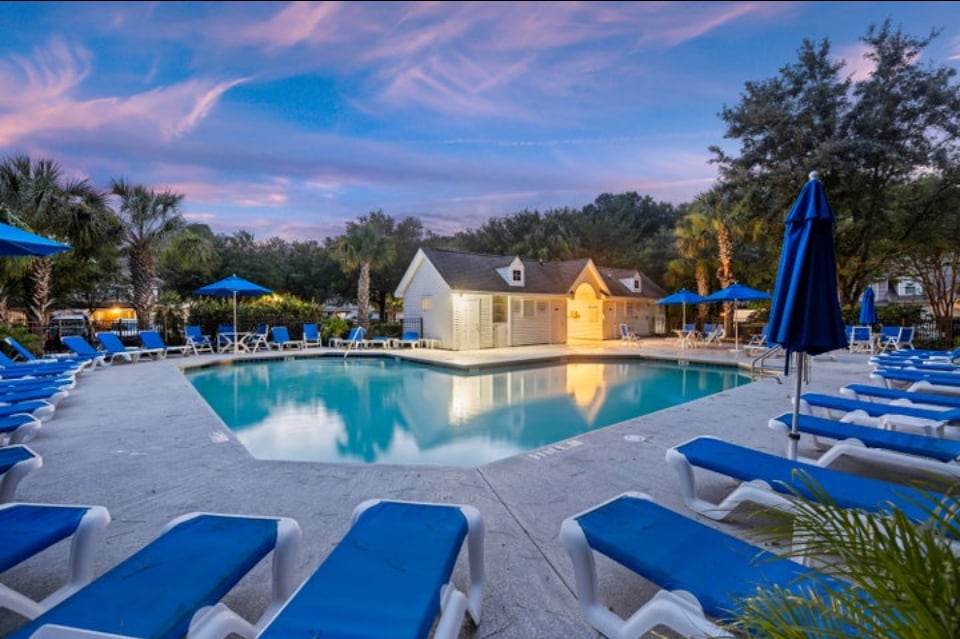 A serene pool area is surrounded by lounge chairs under blue umbrellas, creating a relaxing atmosphere. The pool reflects the evening sky, and a charming white building is positioned nearby amidst lush greenery and palm trees.
