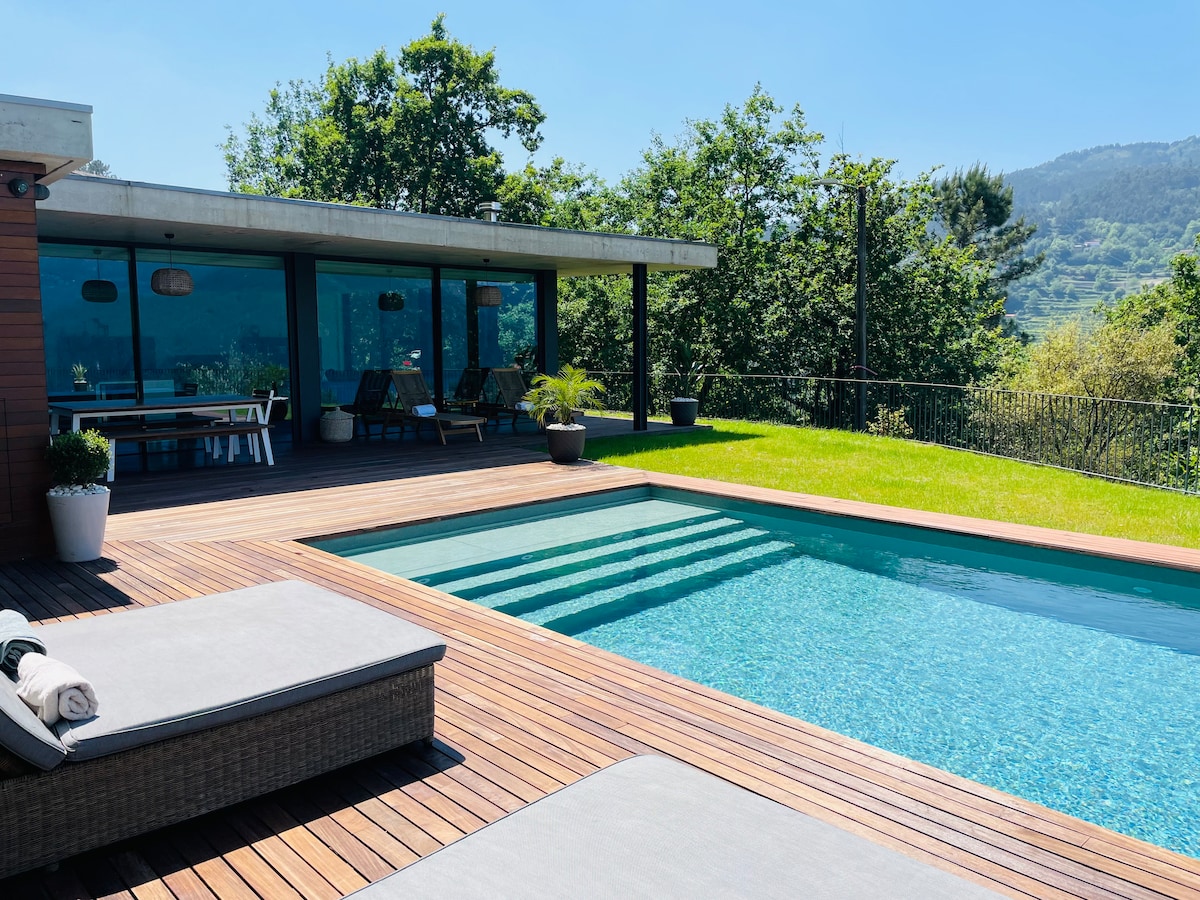 A spacious outdoor area features a private swimming pool surrounded by wooden decking. Sun loungers are positioned nearby, and the lush greenery of the garden provides a serene backdrop. Large glass doors lead into the interior space, inviting natural light.