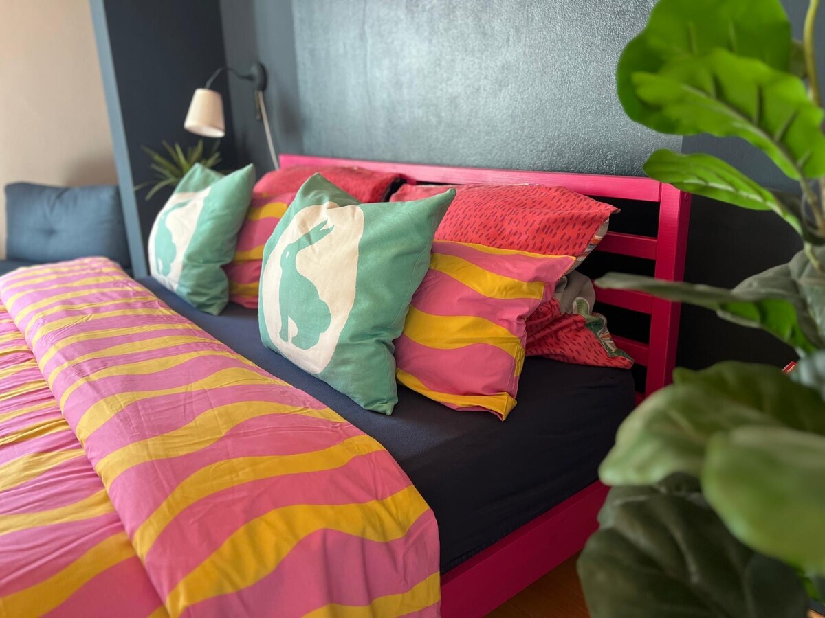 A vibrant bedroom features a spacious bed adorned with a colorful striped blanket and decorative pillows. A bedside lamp provides warm lighting, while a leafy plant adds a touch of greenery to the setting. The wall behind the bed is painted in a dark hue.