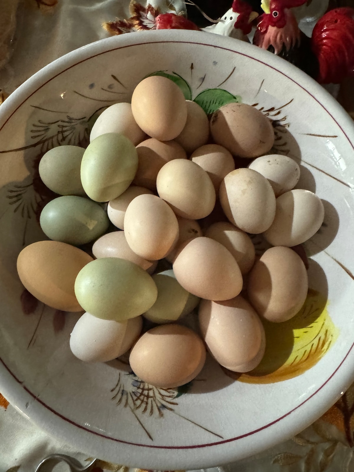 A decorative bowl filled with a variety of eggs in different colors, including pale brown, greenish, and white. The bowl is placed on a patterned tablecloth, adding a charming touch to the presentation. Natural light highlights the smooth surfaces of the eggs.