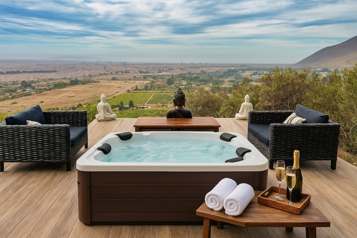 A serene outdoor space is highlighted by a hot tub, surrounded by comfortable seating. A panoramic view of the landscape extends towards the horizon, with mountains in the distance. Two statues are placed nearby, creating a tranquil atmosphere for relaxation.