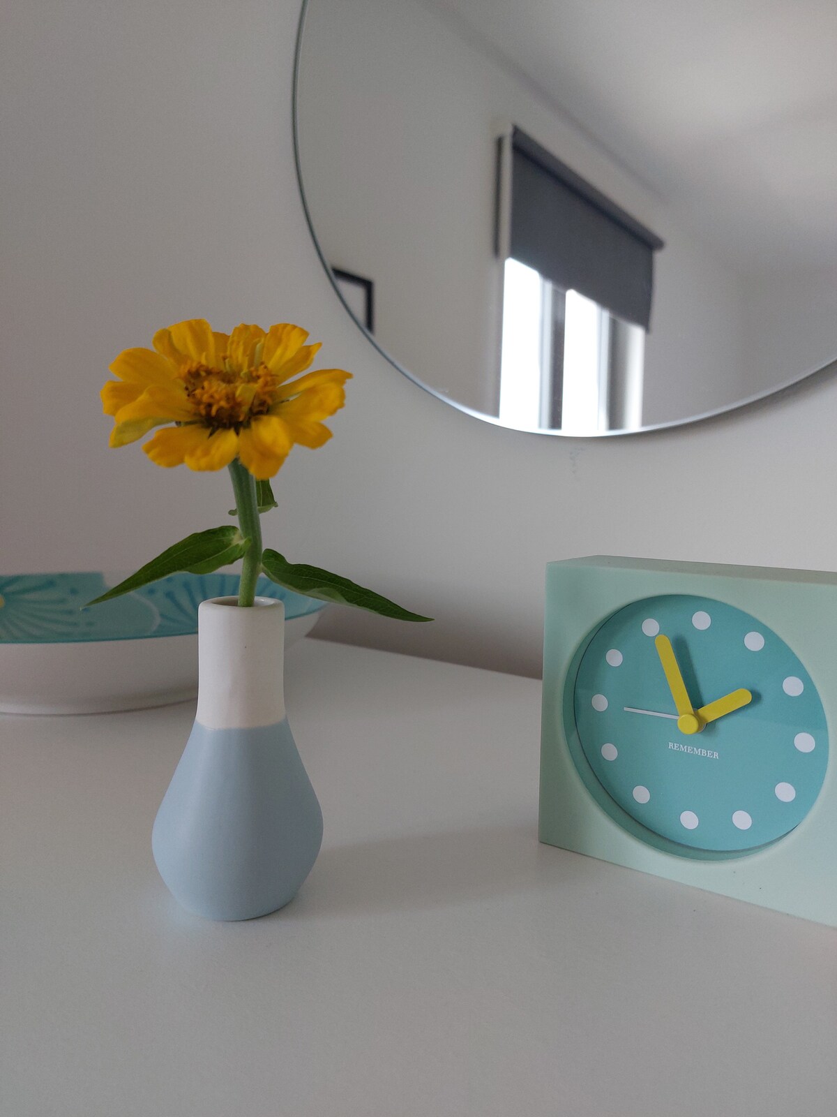 A simple table arrangement is showcased with a small vase holding a yellow flower. Next to it, a round wall clock emphasizes a light blue color with white dots. A mirror reflects softly in the background, adding depth to the bright, modern space.
