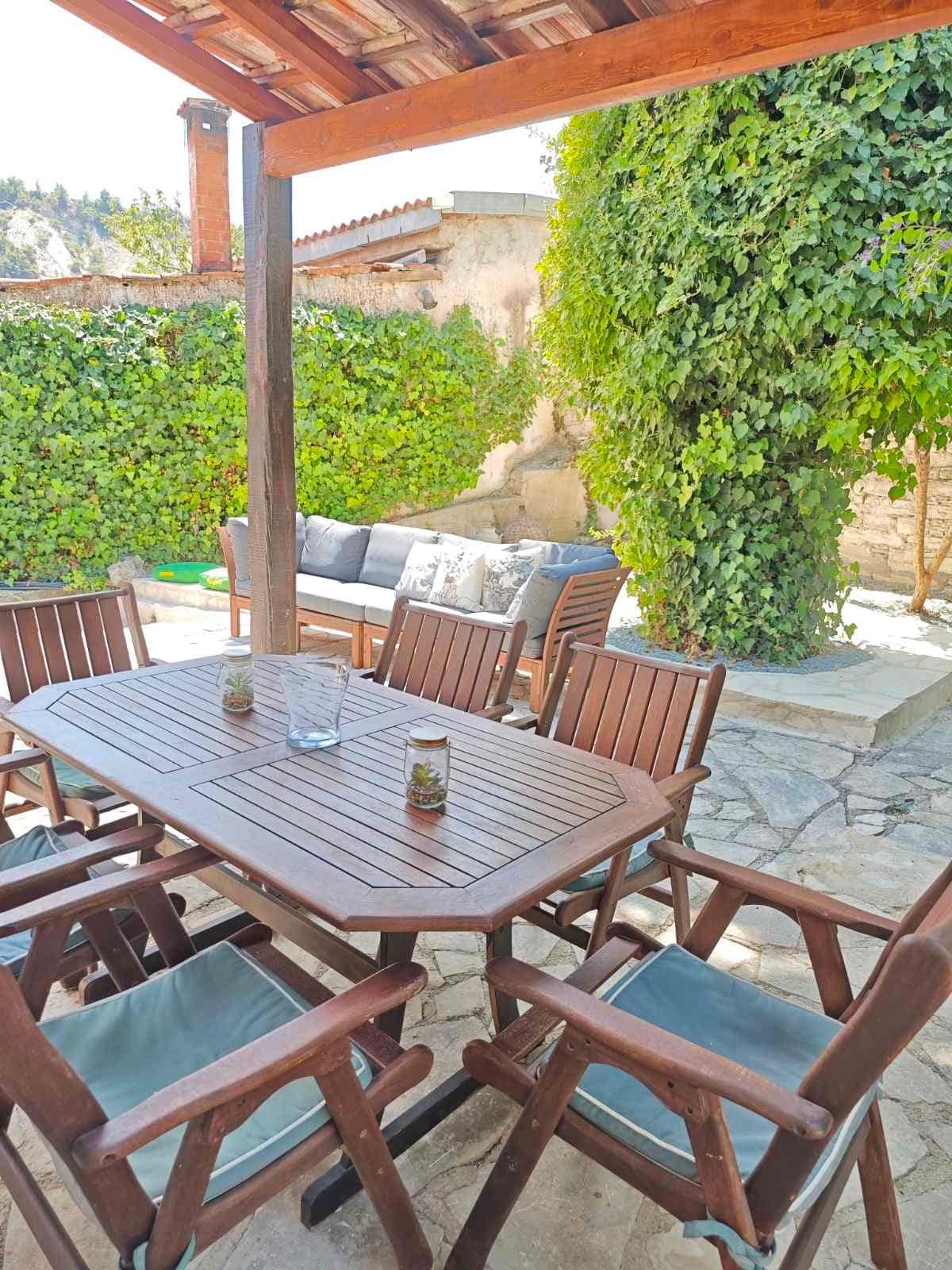 A shaded outdoor dining area features a wooden table surrounded by four sturdy chairs. The space is complemented by a comfortable seating arrangement and is enclosed by greenery, providing a sense of privacy. A stone-paved surface adds to the rustic charm.