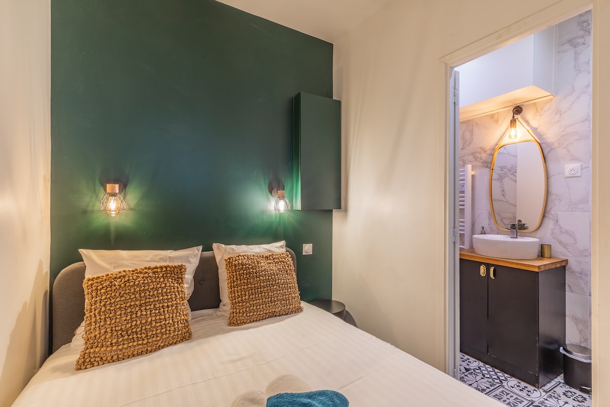 A double bed is positioned against a green wall, featuring two bedside lights. Soft pillows with textured covers are placed on the bed. A doorway leads to a bathroom with marble accents, a circular mirror, and a wooden vanity.