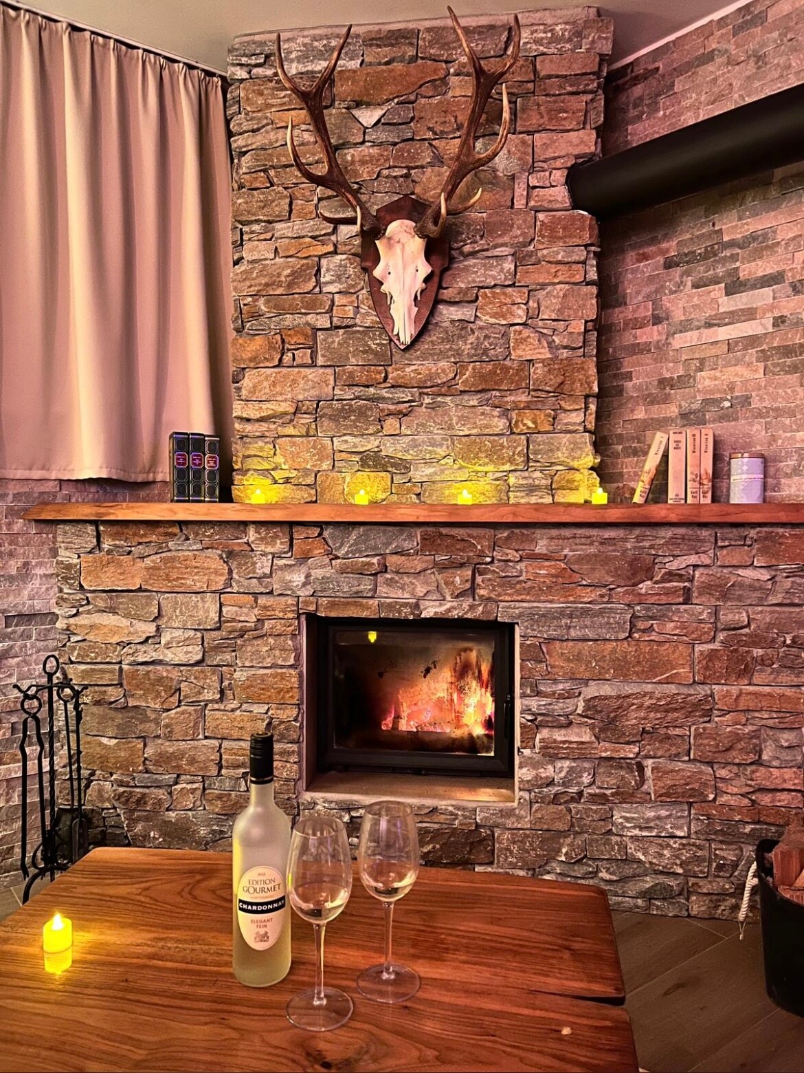 A stone fireplace is showcased with a warm flame, accompanied by a wooden mantel featuring books and decorative items. A rustic wooden table is set with two glasses and a bottle of wine, while soft lighting enhances the serene ambiance.