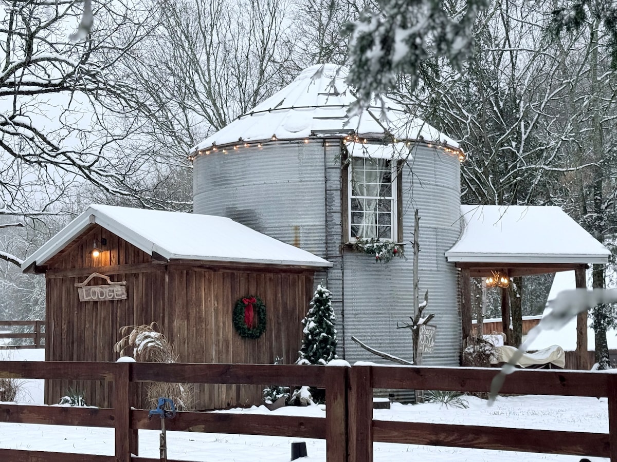 A cozy grain bin structure is partially covered in snow, adorned with warm white string lights along the roof. A rustic wooden building with a festive wreath and a small evergreen tree completes the inviting winter scene, set against a backdrop of snow-covered trees.