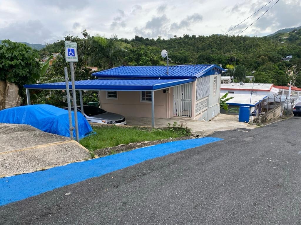 A tiny home is situated on a sloped street, displaying a blue roof and white exterior. A driveway is visible with a parked car, and a blue tarp covers another vehicle. Lush greenery surrounds the home, with hilly terrain in the background.