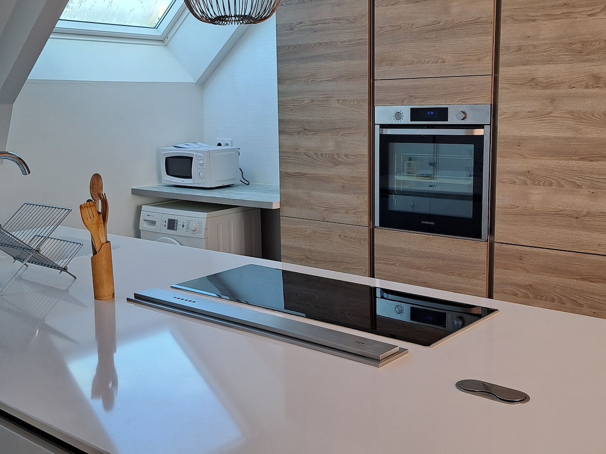 A modern kitchen is showcased with sleek surfaces and warm wooden cabinetry. An induction cooktop is centered on the island, complemented by a microwave and oven built into the cabinetry. Natural light is allowed in through a skylight, enhancing the airy atmosphere.