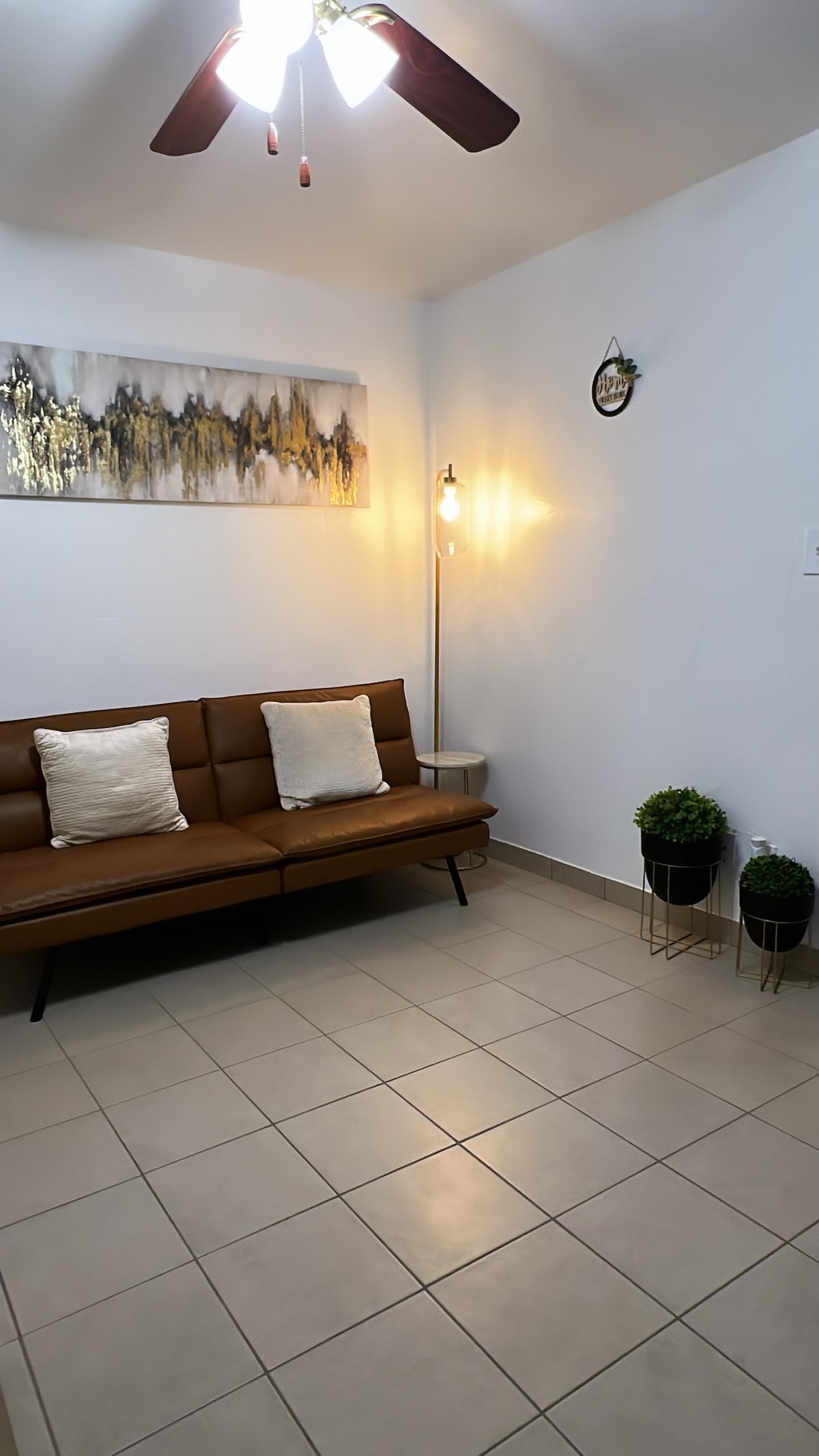 A cozy seating area is featured with a brown sofa complemented by two decorative pillows. A stylish floor lamp provides illumination beside the sofa, and a piece of wall art with abstract designs is displayed above. Potted plants add a touch of greenery.