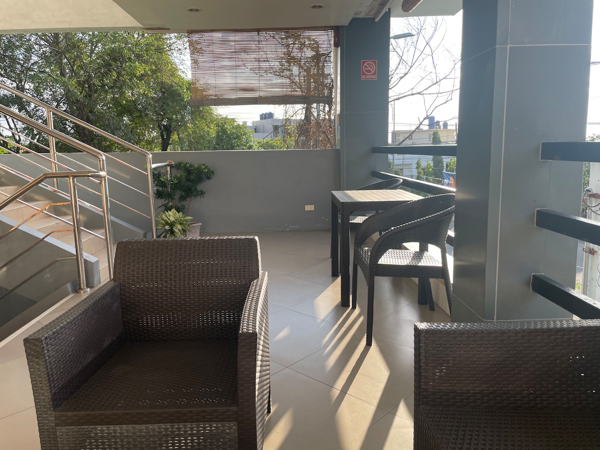 An outdoor seating area features two woven chairs and a small table, surrounded by greenery. Sunlight filters through the nearby windows, casting soft shadows across the tiled floor. A staircase leads to an upper level, indicating a multi-level space.