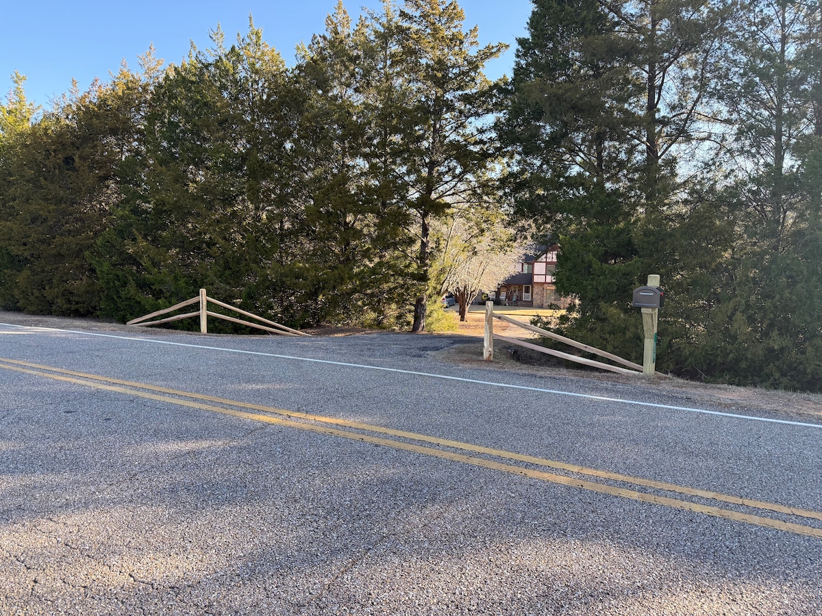 A view of a gravel entrance driveway is seen through wooden gates at the edge of a tree-lined road. The surrounding area is populated with tall evergreen trees, providing a natural border to the property.