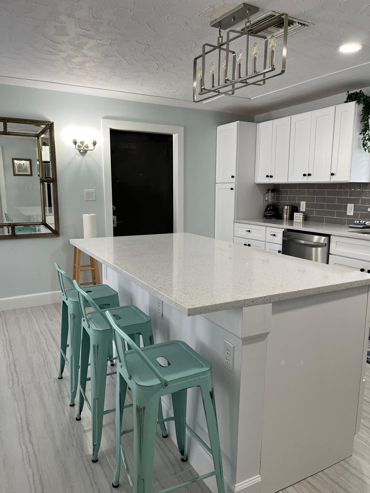 A bright kitchen is presented, featuring a spacious island made of white quartz with three mint-green stools positioned around it. Modern cabinetry in white is complemented by gray backsplash tiles. A large mirror and stylish light fixture enhance the contemporary feel of the space.