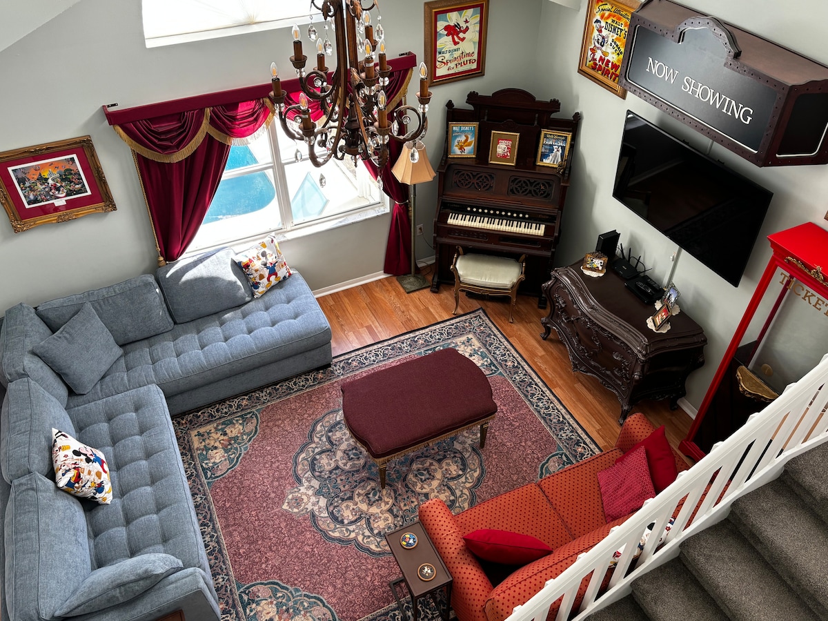 A spacious living area is highlighted by a grand chandelier and features comfortable seating arrangements. A decorative rug adds warmth to the space. A vintage piano and entertainment center are present, complemented by framed Disney artwork and vibrant curtains. Natural light streams through large windows.