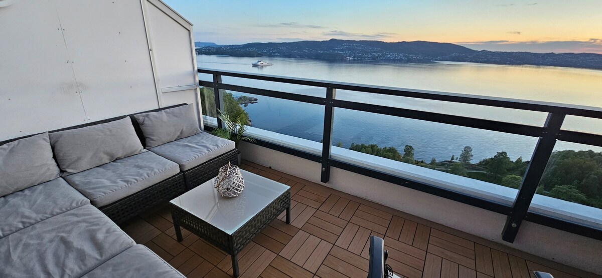 A spacious balcony is featured, furnished with a comfortable seating area and a glass top table. The surrounding railing offers unobstructed views of the water. The deck is laid with wood-patterned flooring, and the scene captures a serene sunset over the landscape.