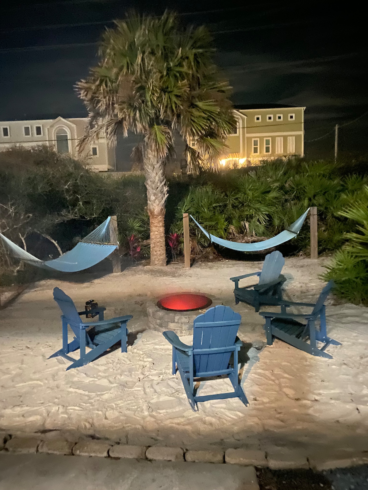 An outdoor relaxation area is depicted, featuring four blue Adirondack chairs arranged around a circular fire pit. Two hammocks are suspended between posts, alongside a palm tree. Soft night lighting enhances the tranquil atmosphere, with greenery surrounding the space.