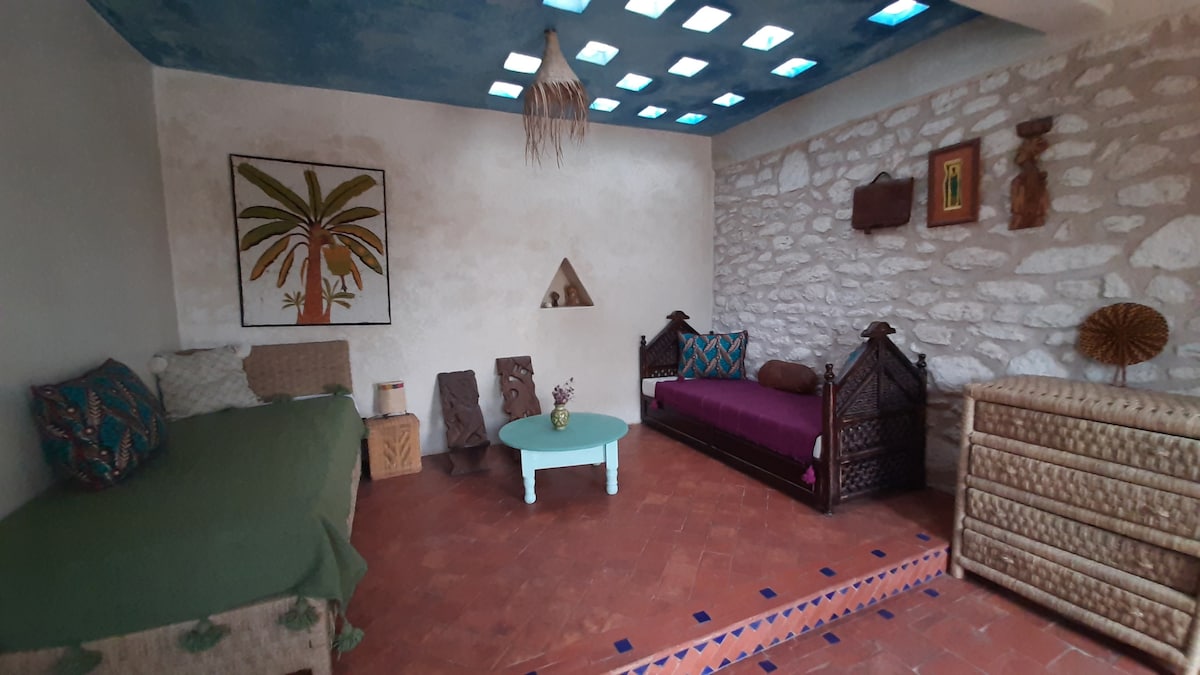 A welcoming living area features a cozy arrangement with two Moroccan style beds and a small round table at the center. Textured walls and a blue ceiling bring a serene feel. Natural light filters through skylights, illuminating decorative elements and local artwork.