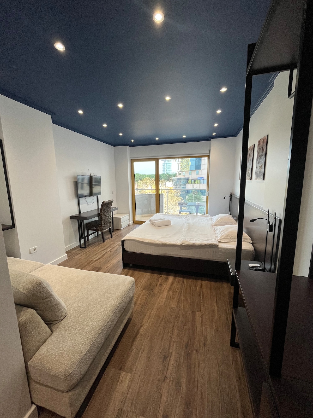 A spacious studio apartment is illuminated by recessed lighting, featuring a chic combination of a comfortable bed and a cozy sofa. A small dining table is visible, with a television mounted on the wall. Large glass doors open to a balcony with views of the outside.