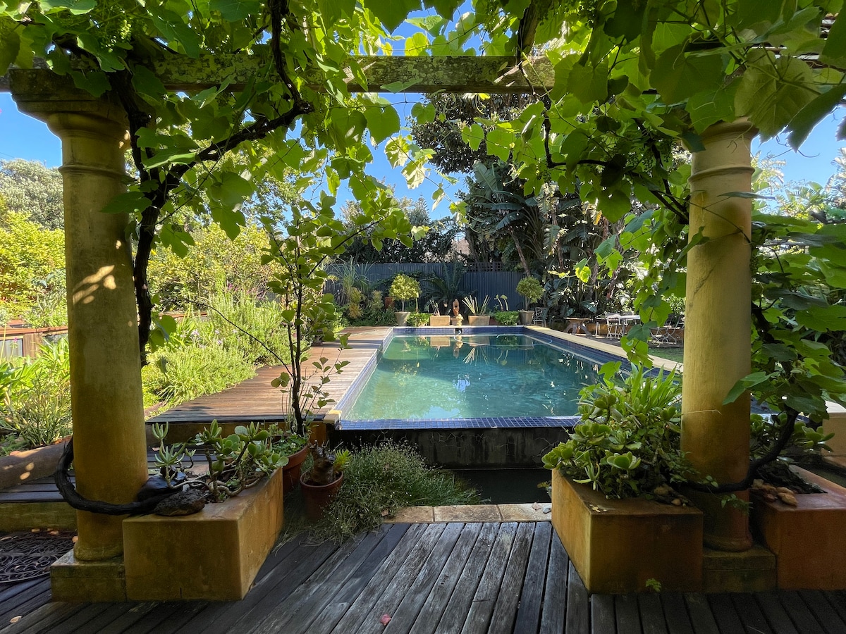 A serene view of a full-size pool framed by lush greenery and architectural columns. Surrounding plants and flowers add natural beauty, while a wooden deck provides a welcoming space for relaxation. The sky is clear, contributing to a tranquil atmosphere.