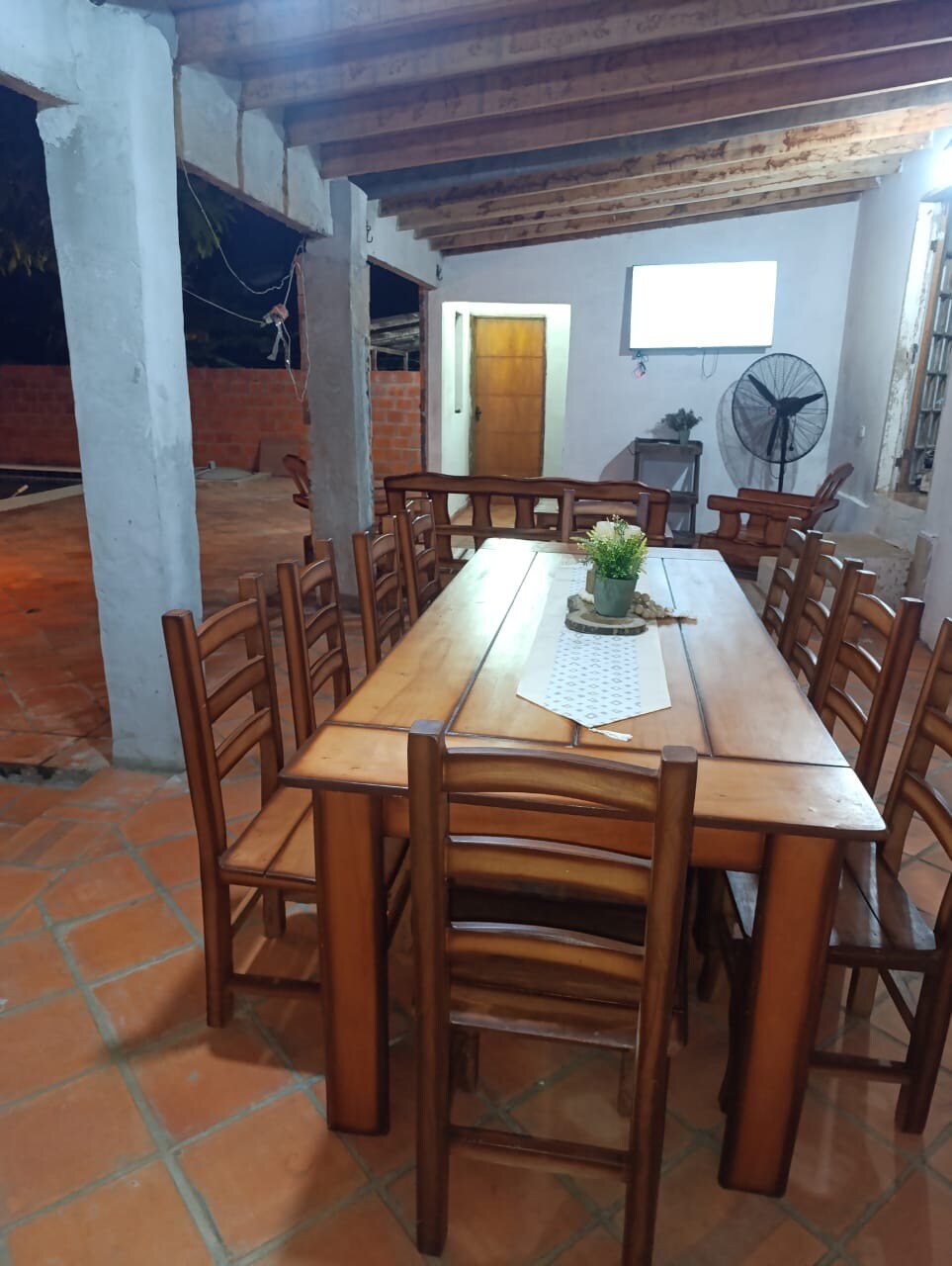 A spacious dining area is furnished with a large wooden table surrounded by thirteen chairs. Exposed wooden beams are visible overhead, complemented by a nearby window. A small plant centerpiece is placed on the table, offering a touch of greenery to the rustic setting.