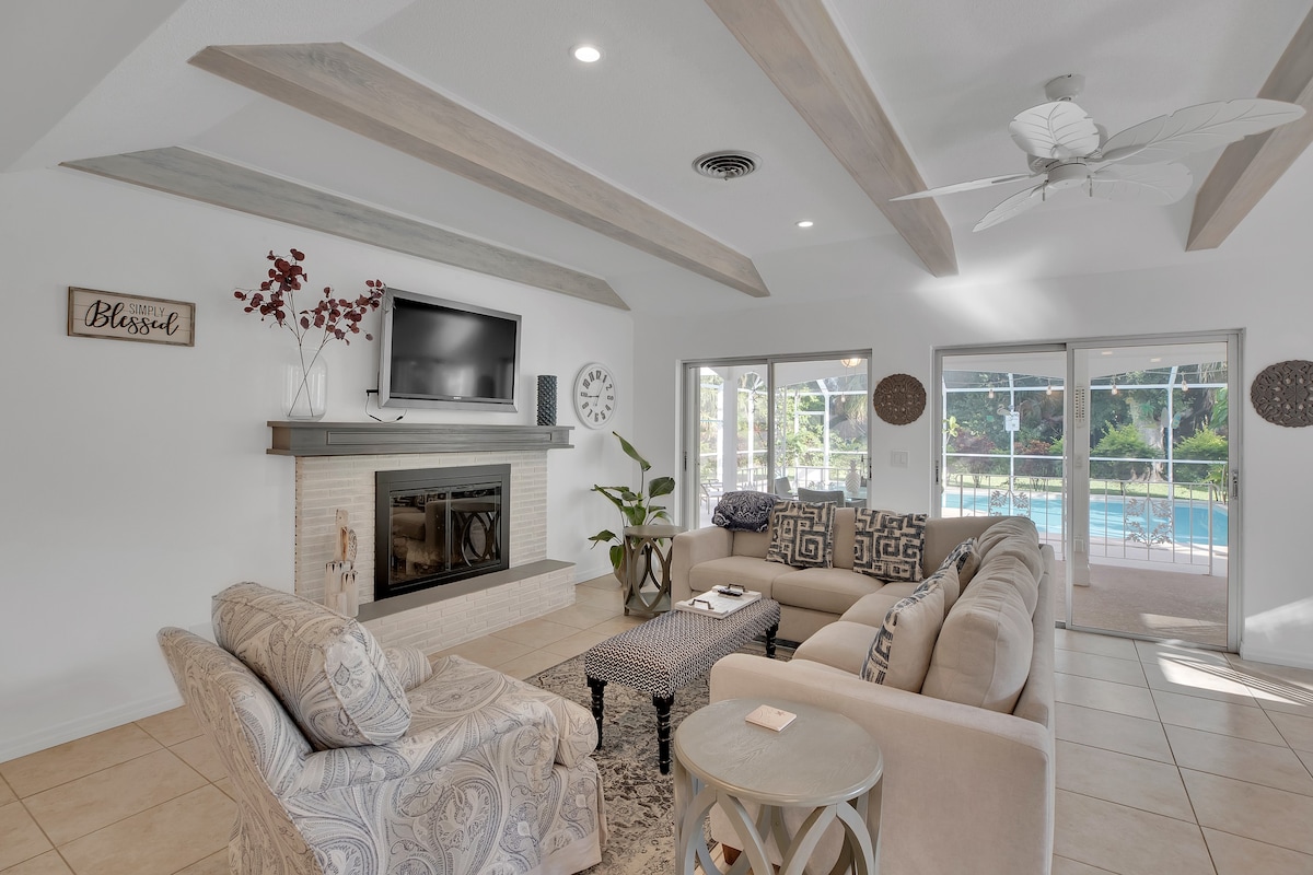 A spacious living area features comfortable seating arranged around a light-colored area rug. A flat-screen television is mounted above a neutral-toned fireplace. Large sliding doors provide a view of the pool area, and natural light fills the space through nearby windows.