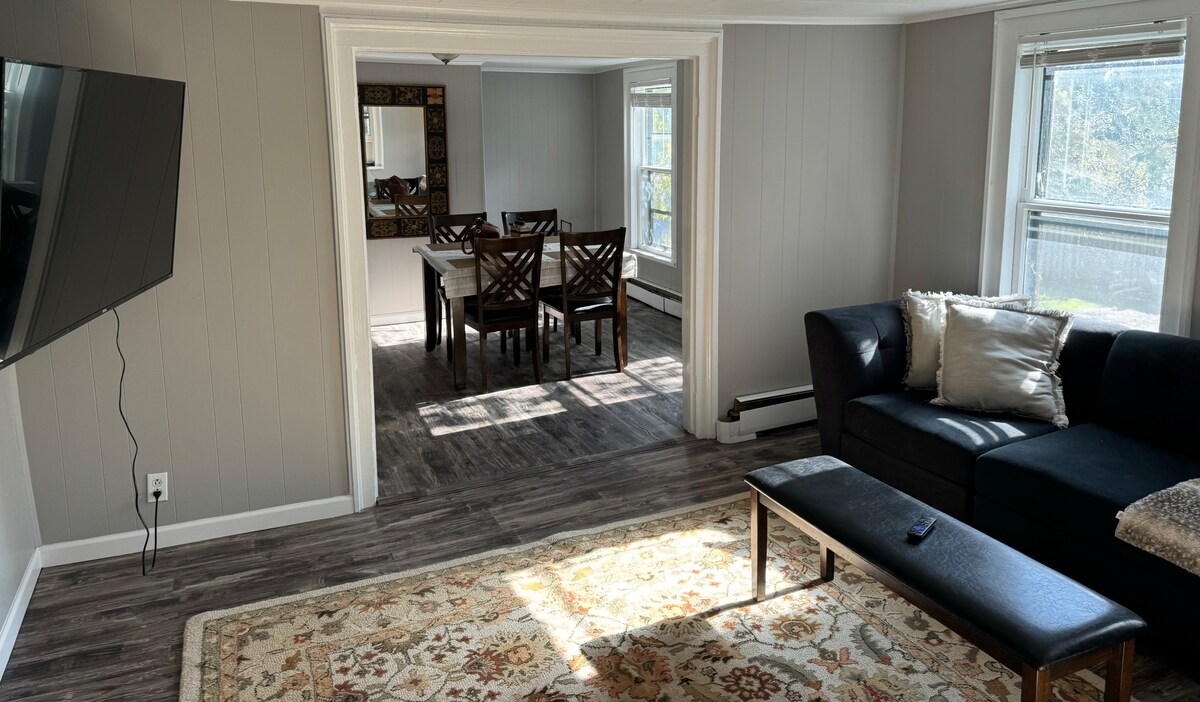A spacious living area features a black couch with decorative cushions and a coffee table positioned on an area rug. A large flat-screen TV is mounted on the wall. Natural light enters through multiple windows, illuminating the dining area with a wooden table and chairs in the background.