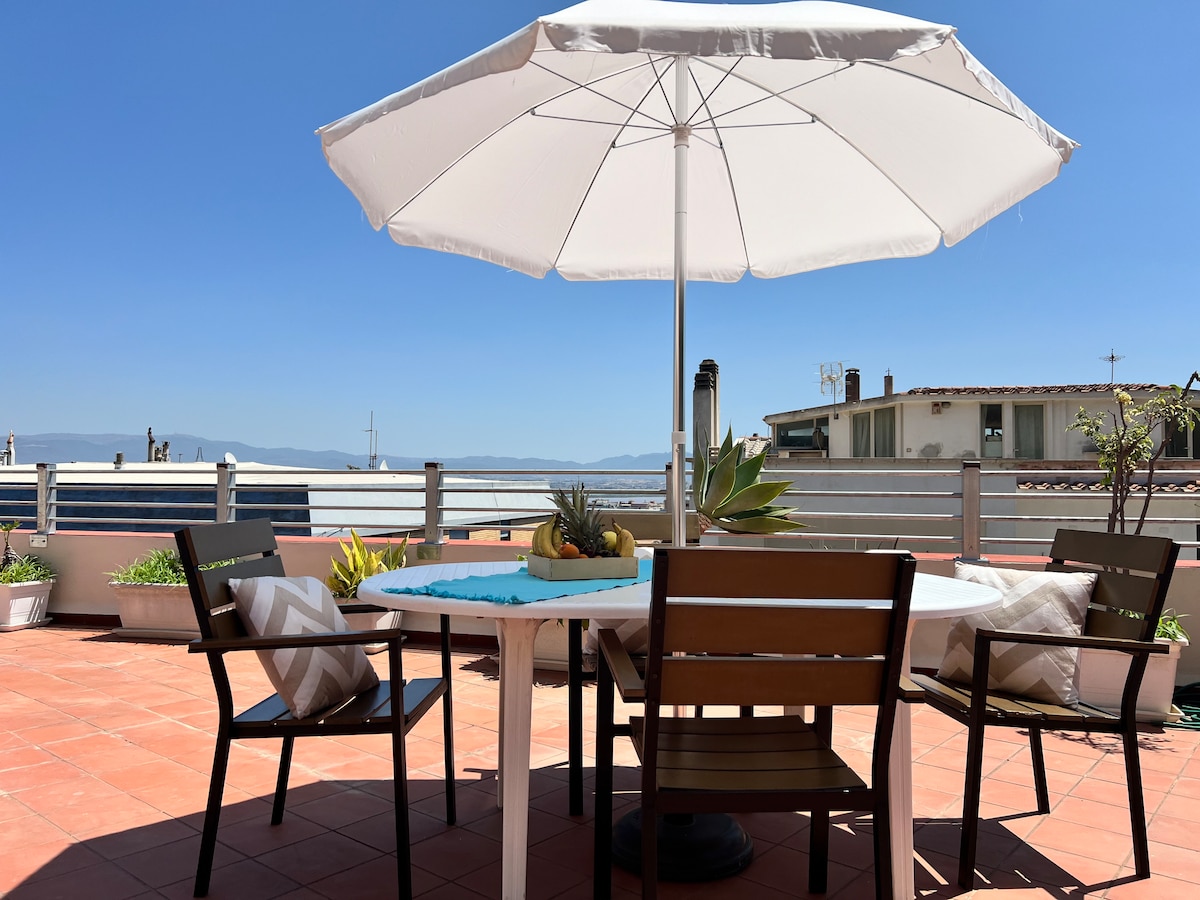 An outdoor terrace is shown, featuring a round table set with a light umbrella and six wooden chairs. Potted plants are arranged around the area, providing a relaxed and inviting environment, perfect for dining or enjoying the views.
