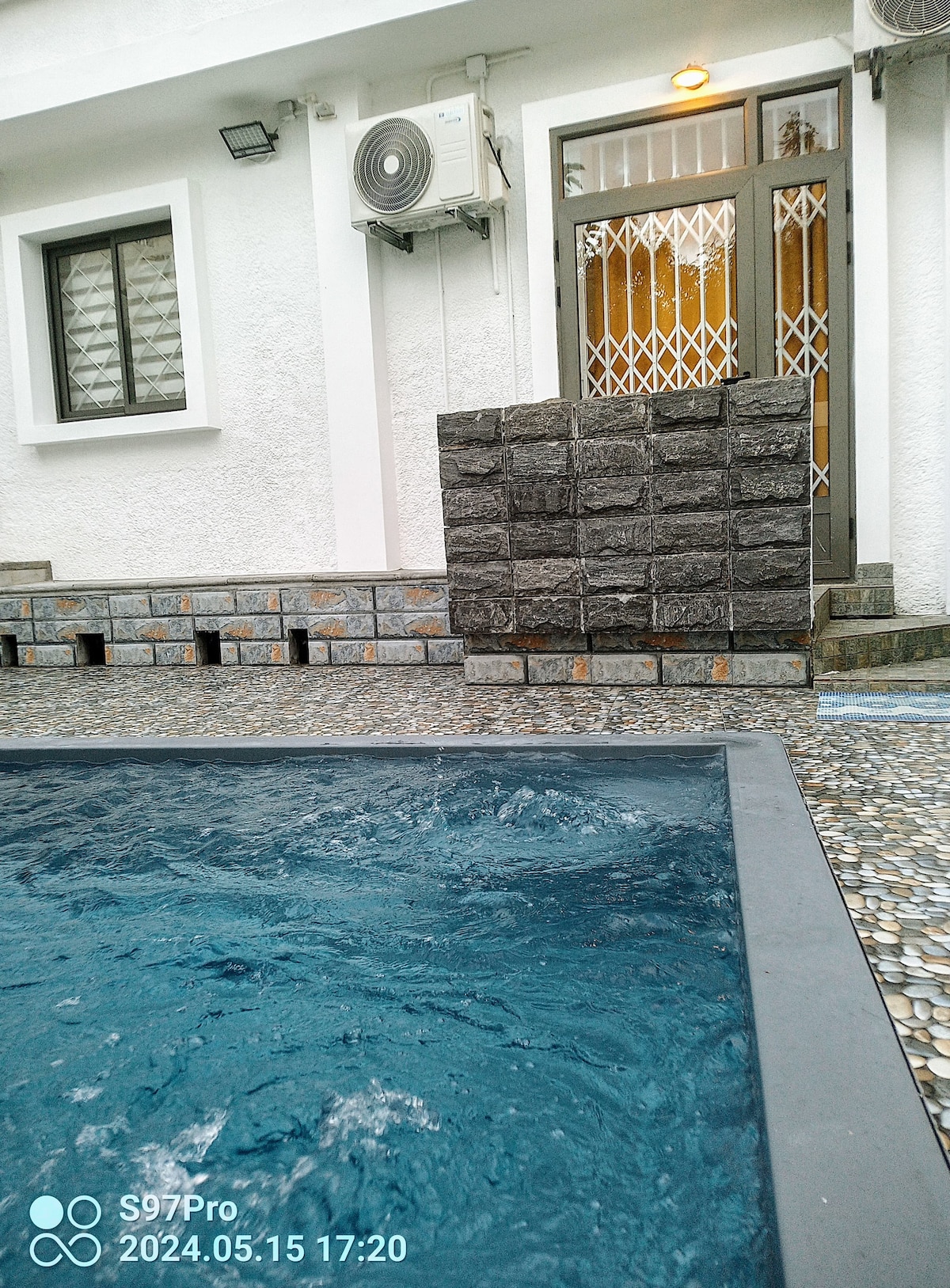 A view of a private pool is captured, with water gently rippling in shades of blue. The surrounding area includes textured stone walls and a nearby entrance featuring a secure door. Air conditioning units are visible on the exterior wall of the villa.