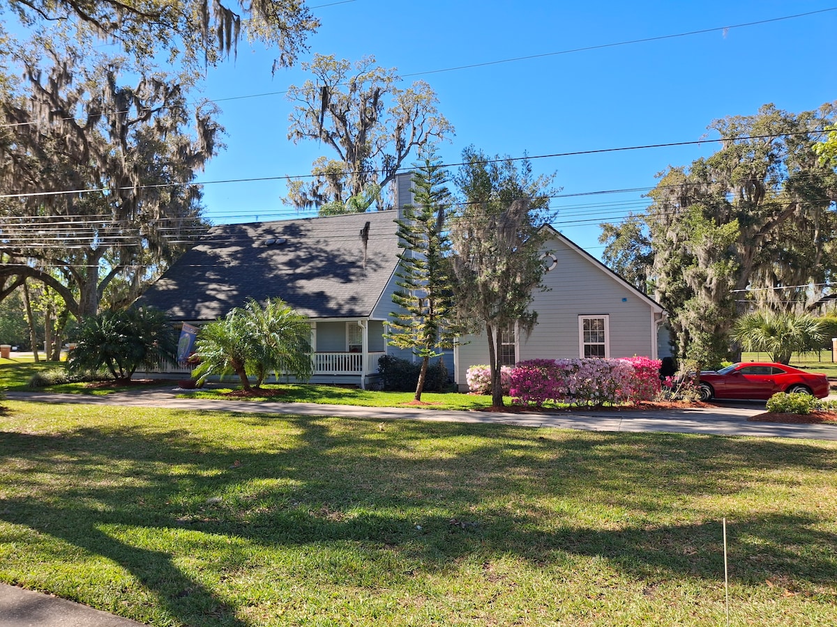 A charming two-story house is situated on a spacious lawn, surrounded by lush greenery and flowering shrubs. Large trees provide shade, and a vibrant red vehicle is parked on the driveway. A porch is visible at the front, inviting a sense of relaxation.