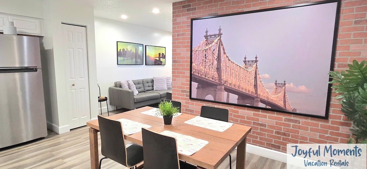 A dining area is presented with a wooden table set for four, surrounded by simple black chairs. A large wall-mounted TV displays an image of a bridge, while a light gray sofa sits in the background, accompanied by framed artwork. A refrigerator is visible to one side.