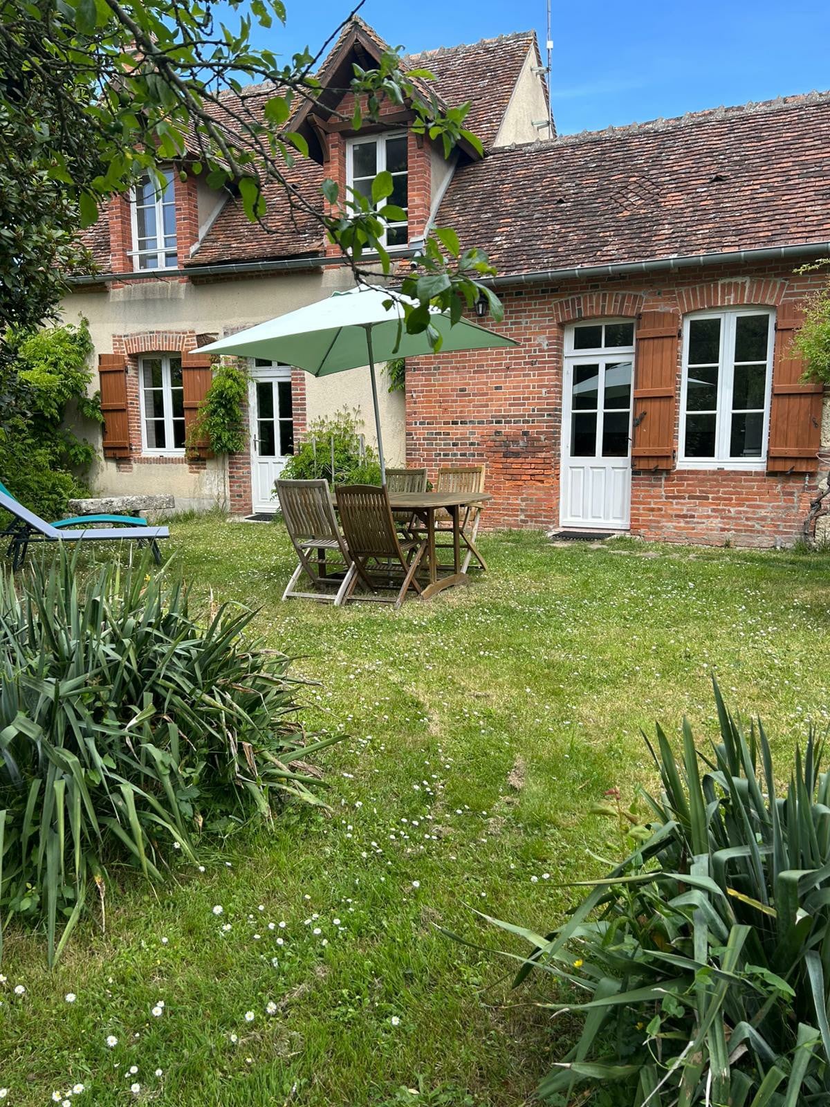 A serene garden space features a wooden table surrounded by chairs under a large umbrella. The lush green lawn is bordered by flowering plants and shrubs, alongside a brick house showcasing traditional architecture with wooden shutters. Bright sunlight highlights the peaceful setting.