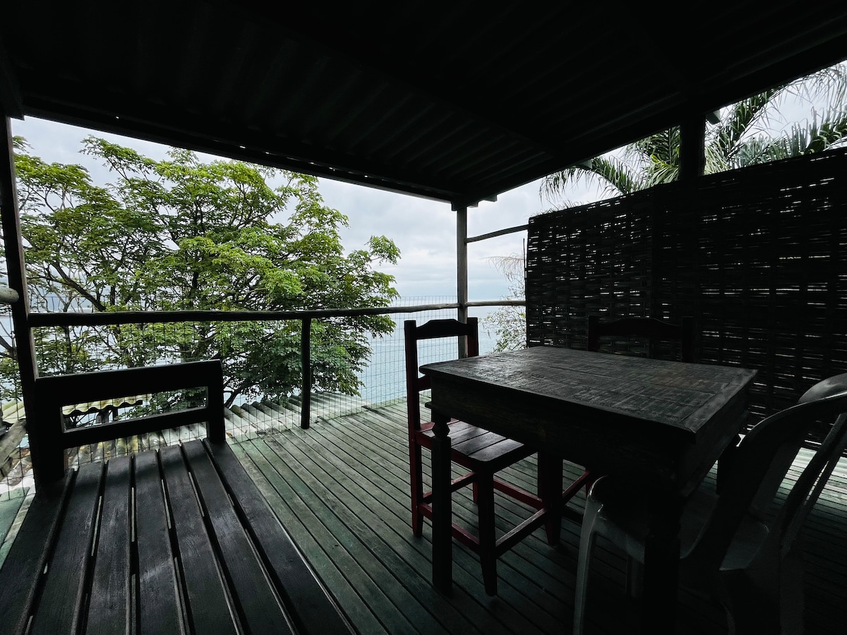 An outdoor seating area is visible on a covered balcony, featuring a wooden table surrounded by chairs. Lush greenery is seen in the background, with the ocean stretching out beyond. A railing provides safety, enhancing the sense of privacy in this tranquil space.