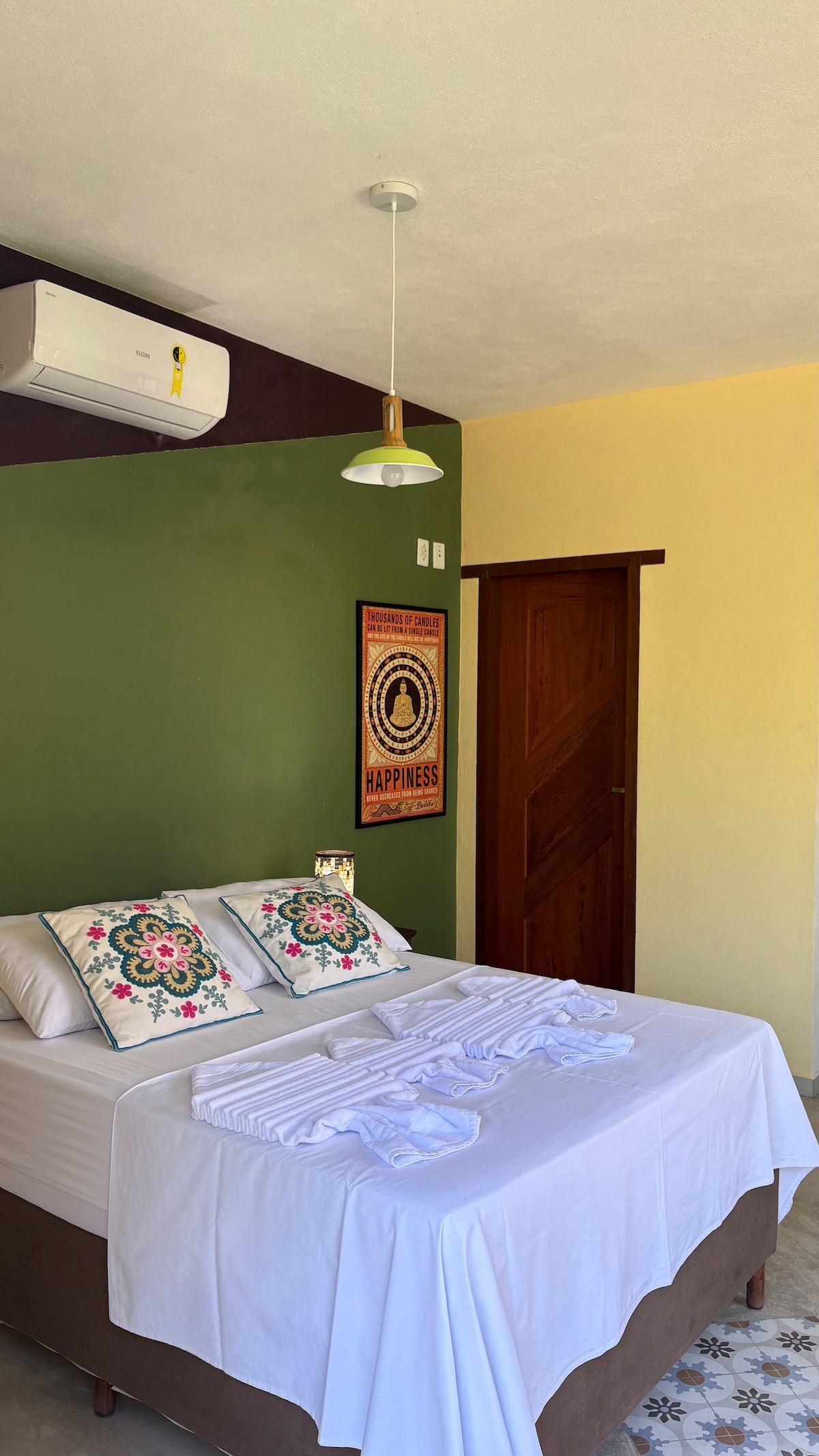 A spacious bedroom is featured with soft white linens on the large bed, adorned with colorful decorative pillows. A wall in a calming green tone enhances the space, while an air conditioning unit is mounted above. Natural light filters through, illuminating the room.