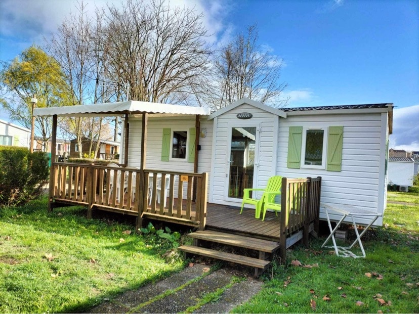 A mobile home is set on a grassy area, featuring a semi-covered terrace with white chairs and a green chair. The exterior showcases light-colored siding and green window shutters, with trees in the background and blue skies above.
