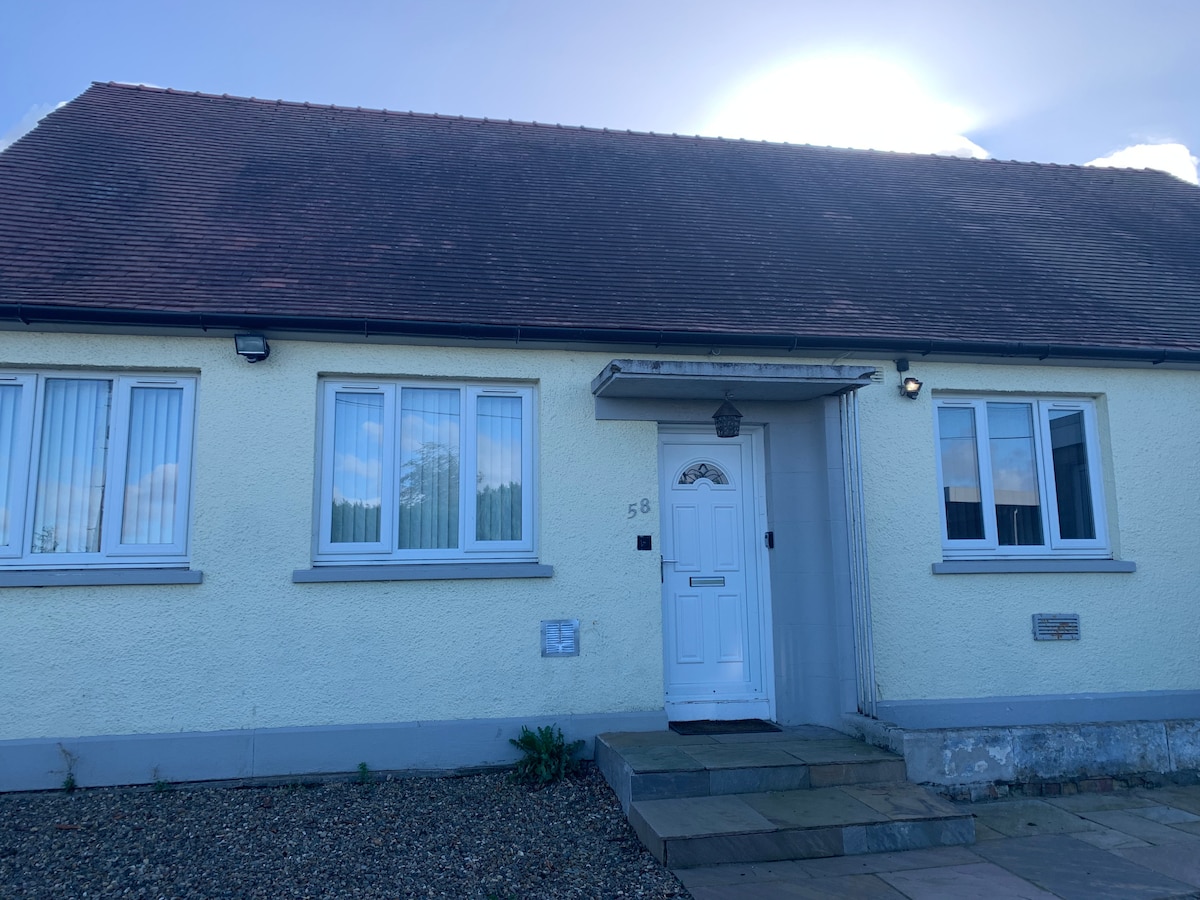 The exterior of a single-story bungalow is presented, featuring a light yellow facade and a sloped roof. Windows are lined symmetrically, allowing ample natural light inside. A stone path leads to the front door, which is centered, enhancing accessibility.