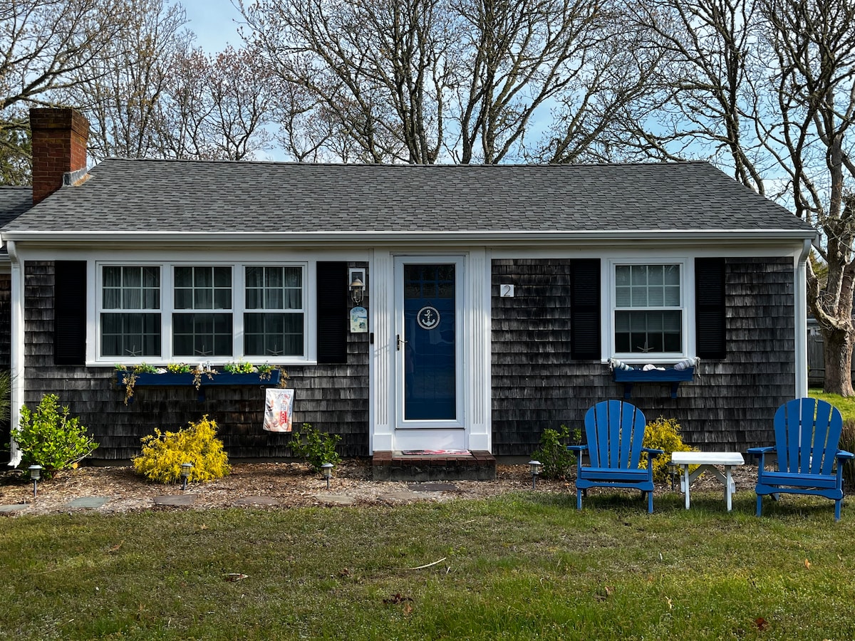 The front exterior of the house features a shingled facade with contrasting dark shutters. Two blue chairs sit in front of the entrance surrounded by small bushes and flowers. A welcoming blue door is centered between multiple windows, enhancing the home's charm.