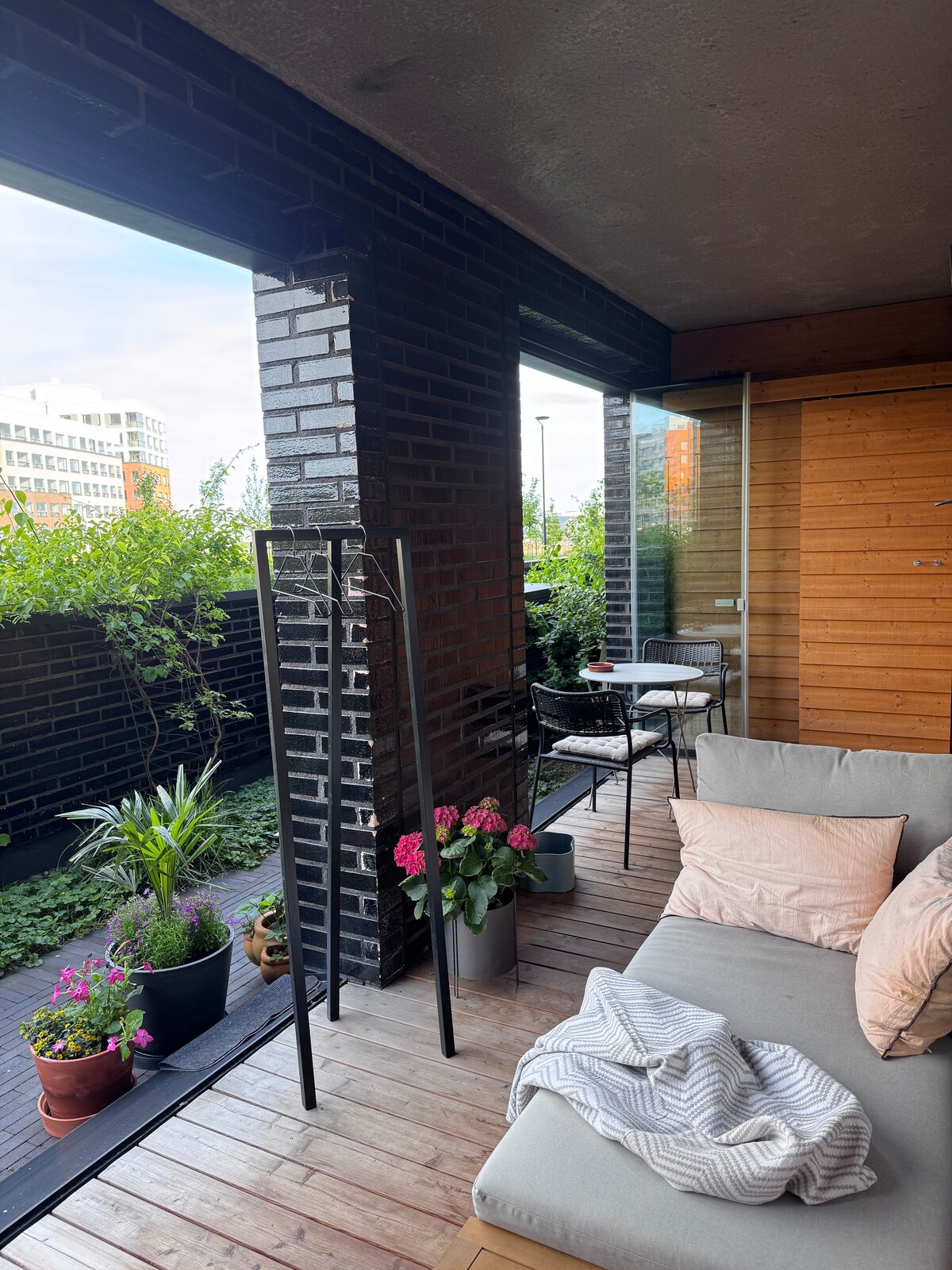 An outdoor balcony with wooden flooring is seen, featuring potted plants and flowers. A comfortable sofa is positioned against one wall, complemented by a light-colored blanket. A small dining table with chairs is visible in the background, surrounded by greenery.