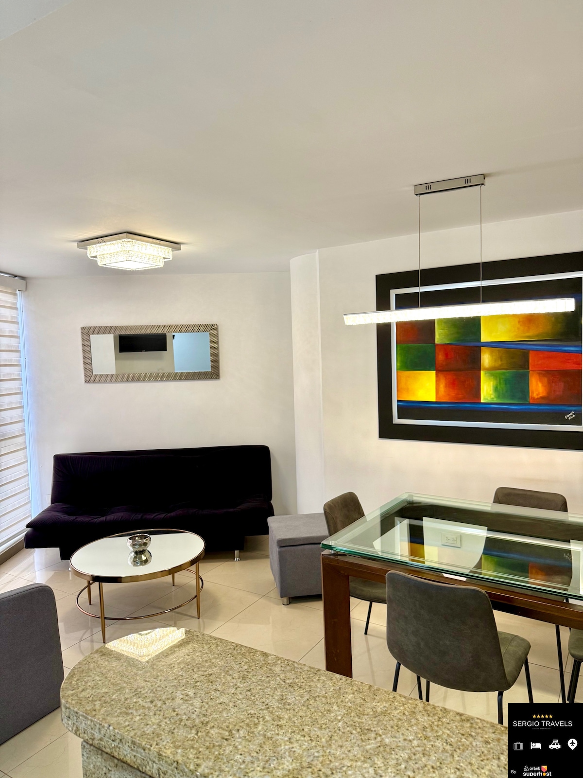 A modern living and dining area is showcased, featuring a comfortable sofa, a glass table framed by four stylish chairs, and a contemporary chandelier. A colorful abstract painting adorns the wall, while natural light enters through large windows covered with light blinds.