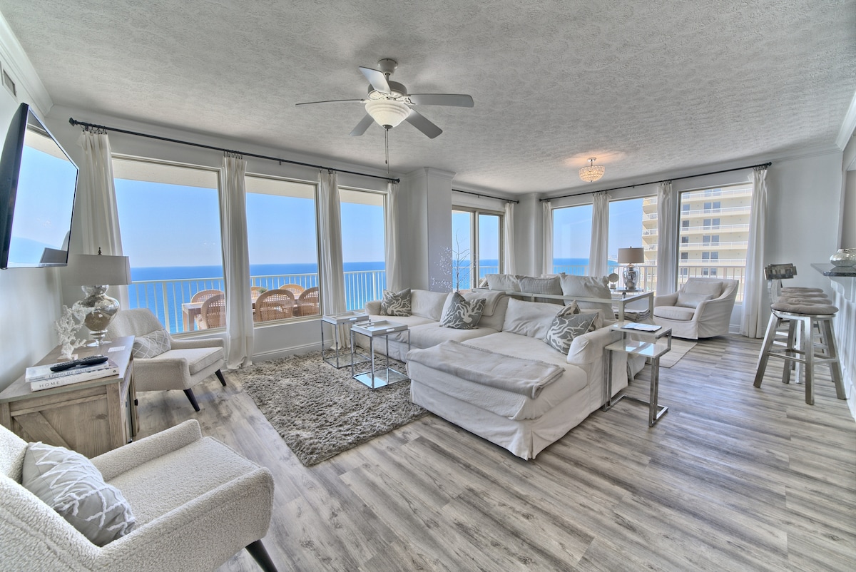 A spacious living area features light-colored furnishings, including a sectional sofa and accent chairs, with large windows showcasing beach and ocean views. A soft area rug and a ceiling fan enhance the comfort, while a flat-screen TV is mounted on the wall.