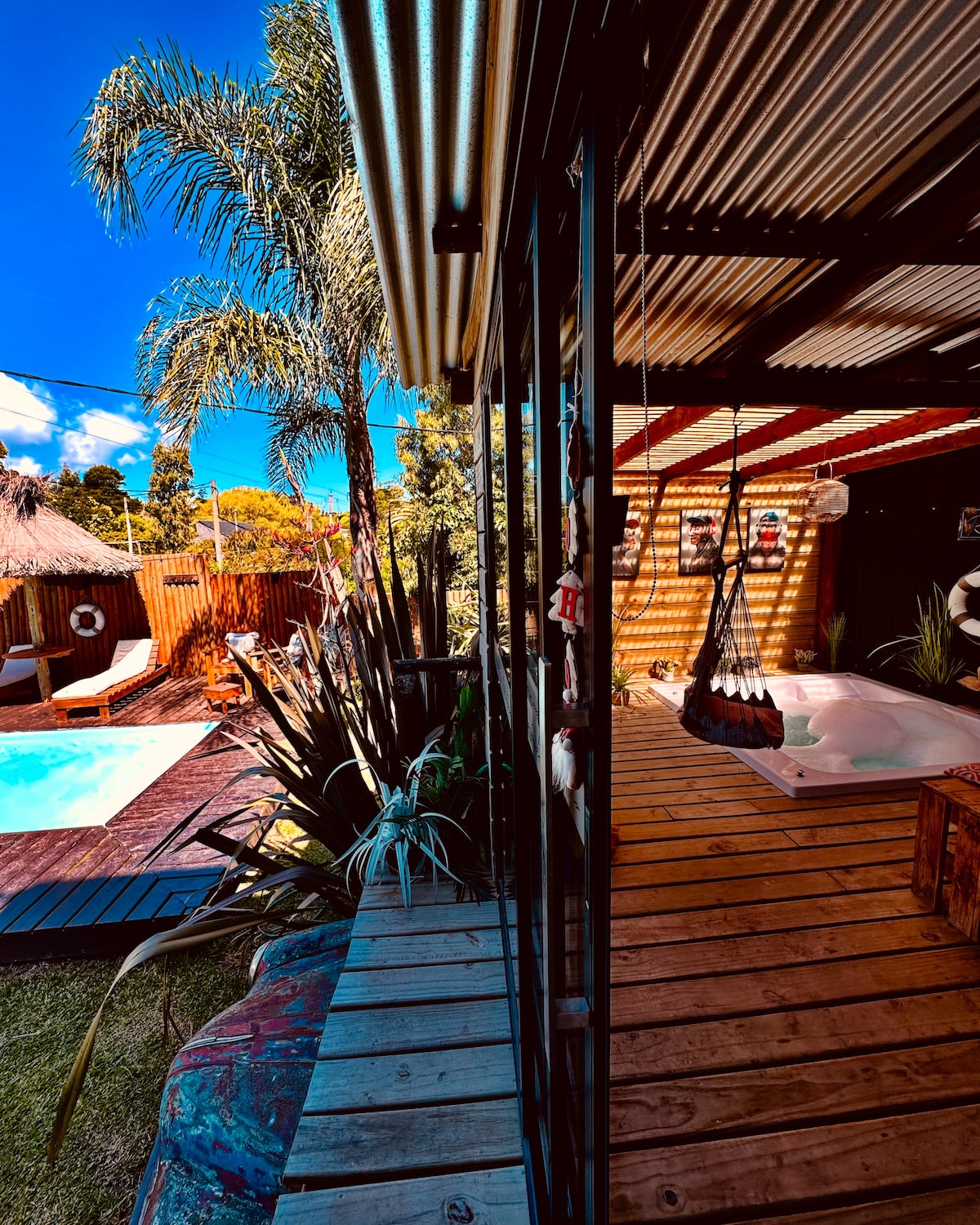 An exterior view captures a private outdoor area featuring a swimming pool and a heated jacuzzi. Lush greenery and tropical plants surround the space. A hammock hangs nearby, while sun loungers are positioned on wooden decking, inviting relaxation under a clear blue sky.