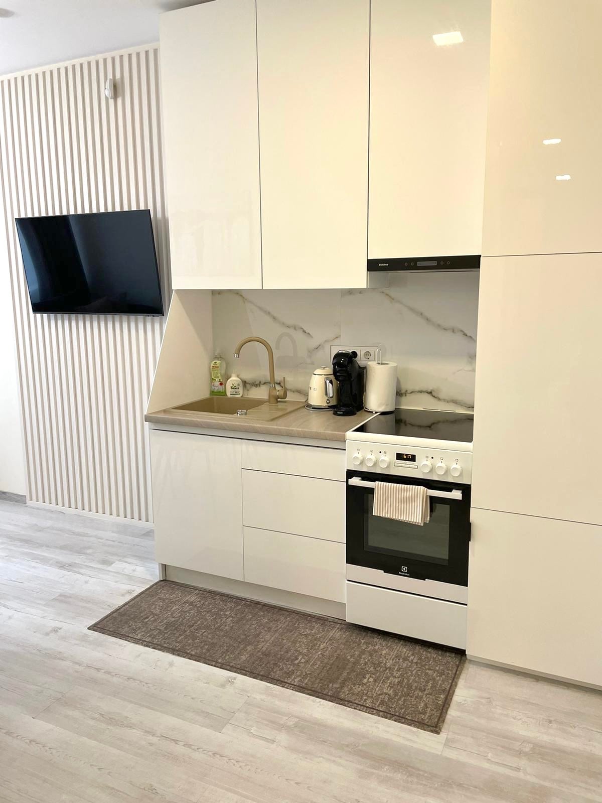 The kitchen features sleek white cabinetry and a light-colored countertop, with a stove and oven visible. A wall-mounted television is positioned above the countertop, and small appliances are neatly arranged. A rug lies on the floor, adding a touch of comfort to the space.